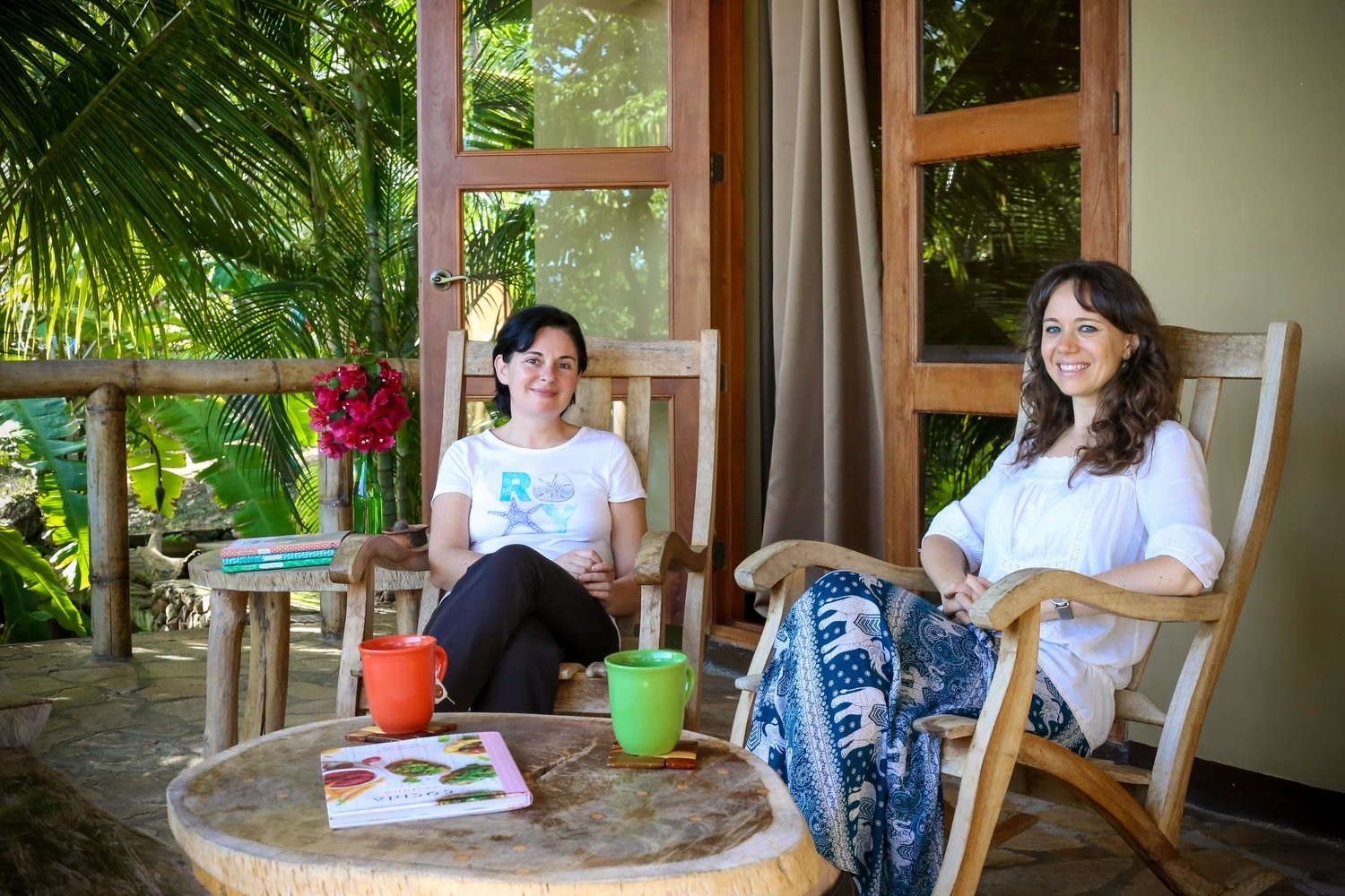 Two women sitting in wooden rocking chairs on a tropical porch with coffee mugs and books.
