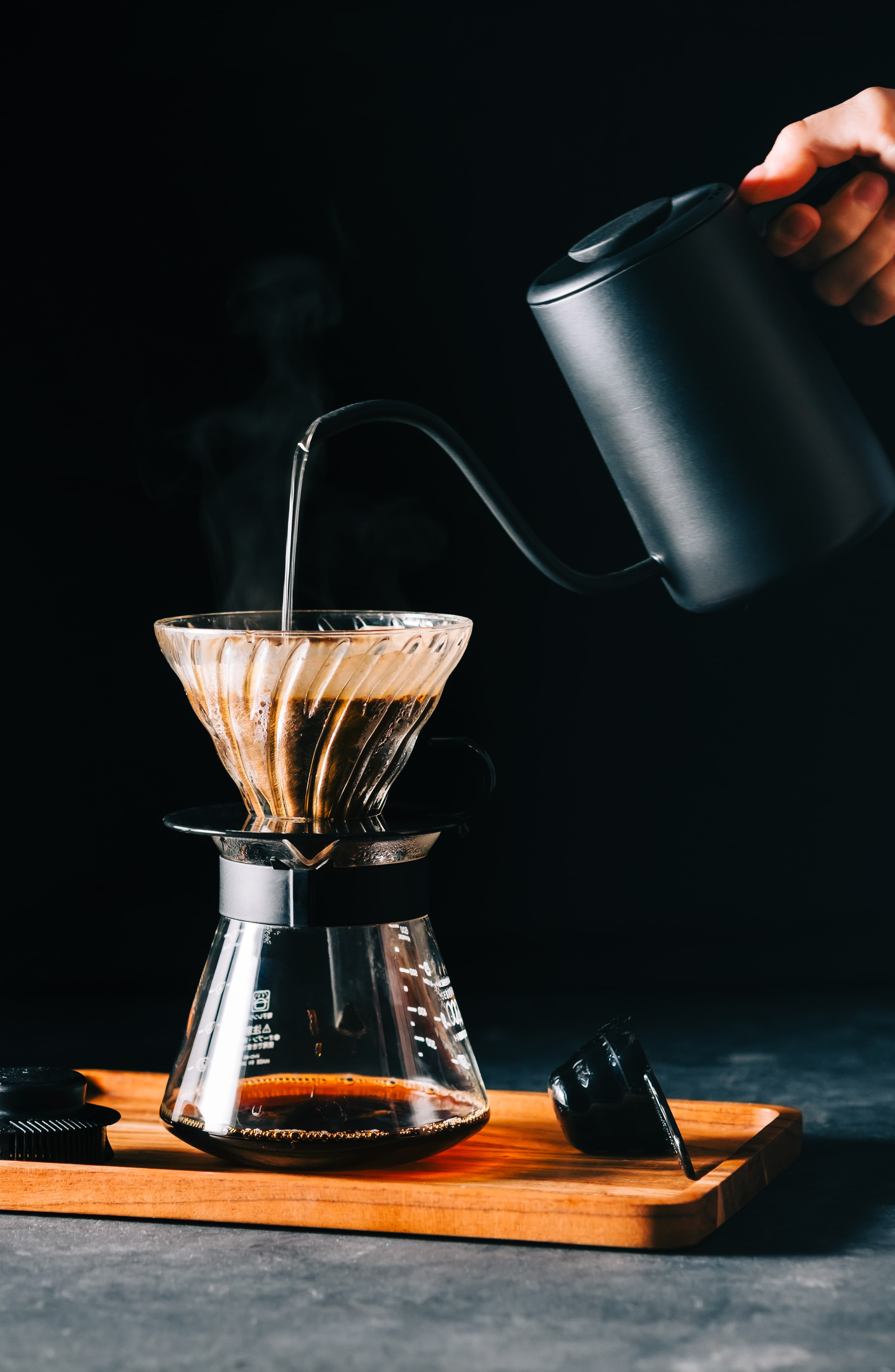 Hand pouring hot water from a gooseneck kettle over a coffee dripper, with a glass carafe below, in a dark, moody setting.