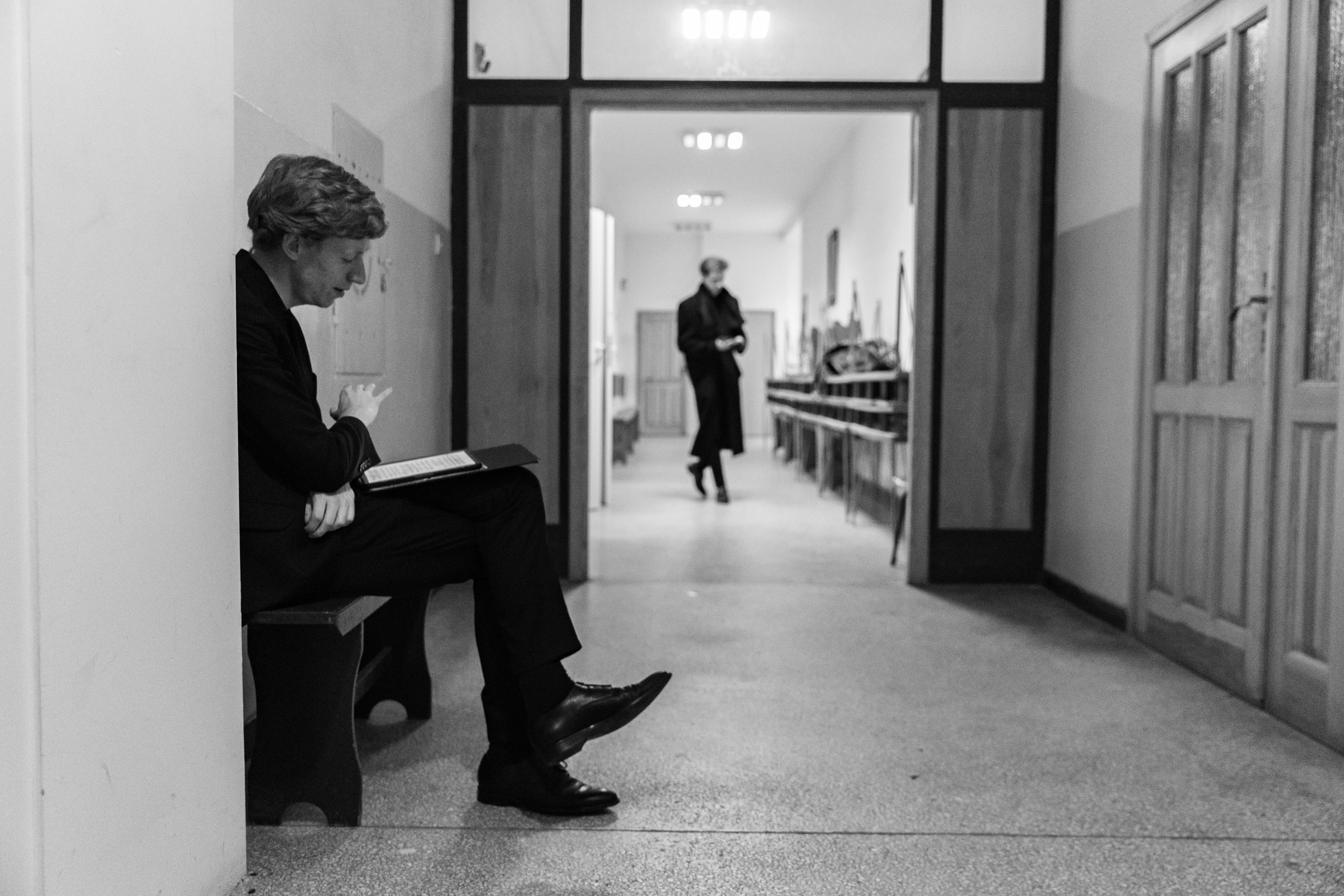 Mężczyzna w garniturze siedzący na ławce z książką w ręku, w korytarzu z widokiem na drugą osobę w czarnym płaszczu idącą w stronę kamery. Korytarz o jasnych ścianach z drzwiami i gzymsem w tle.
