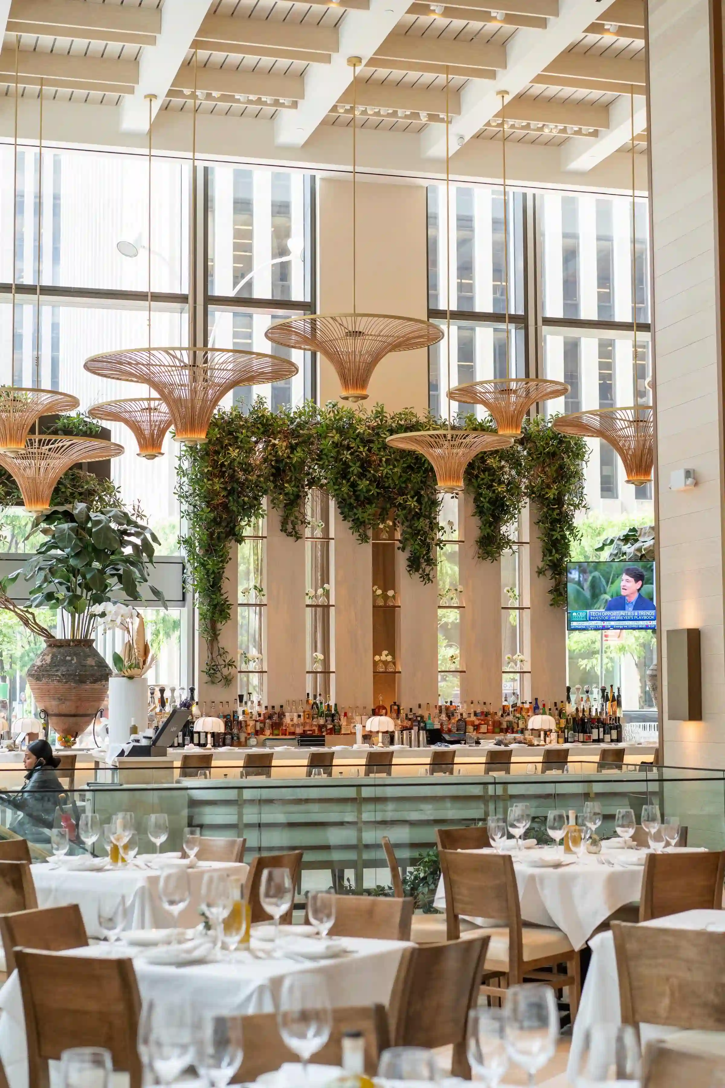 Elegant dining area with tall windows, lush greenery, and tables set for guests. Bright and inviting atmosphere.