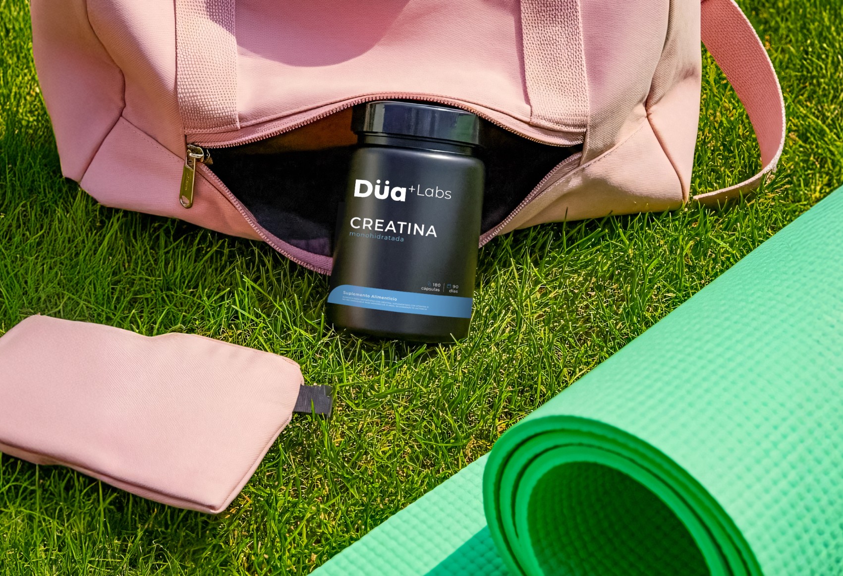 A pink gym bag sits on vibrant green grass, partially unzipped to reveal a container of creatine from Dua Labs, with a green yoga mat rolled up alongside.