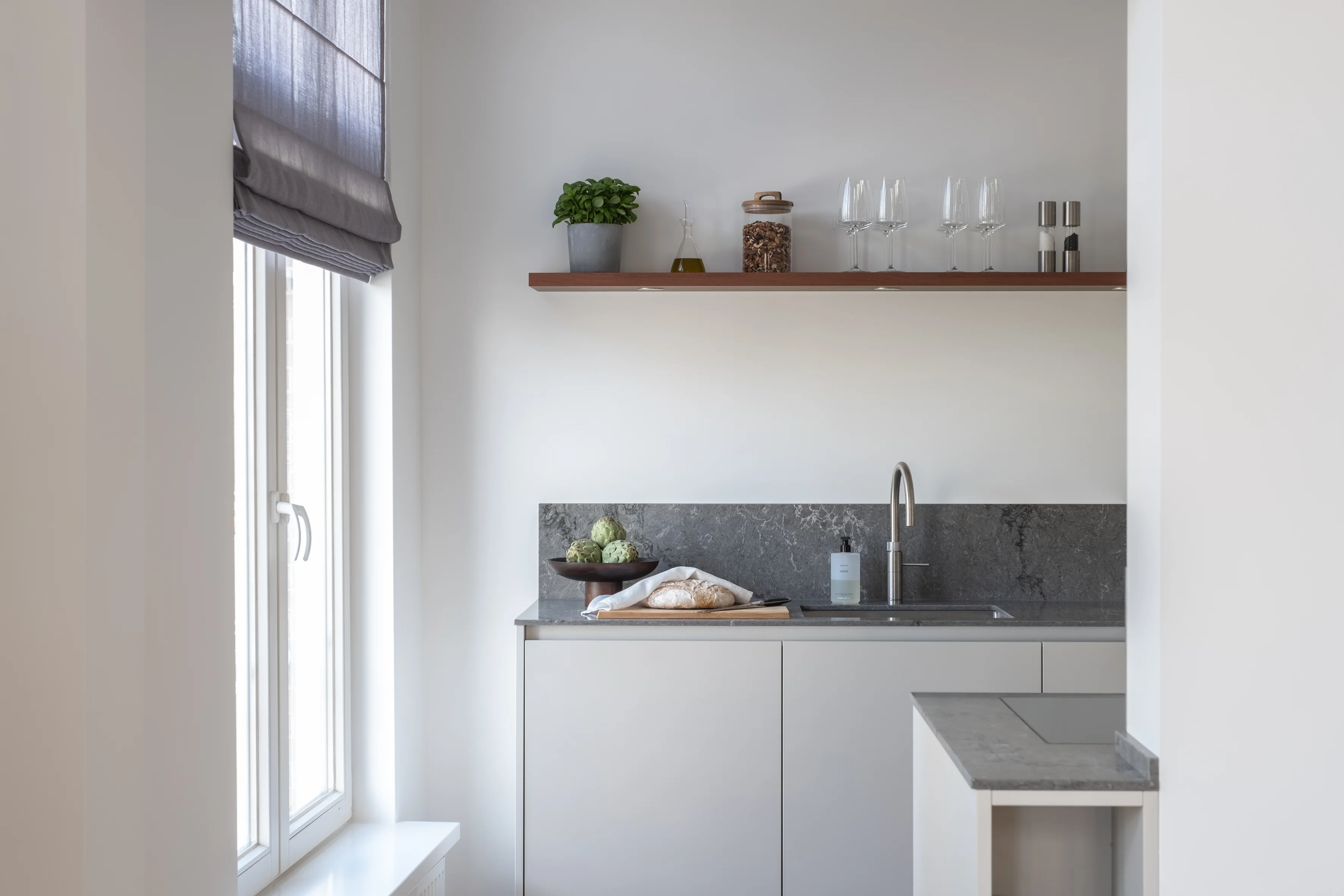 Minimalist kitchen niche with grey stone backsplash, open walnut shelving, glassware, and lightgrey cabinetry