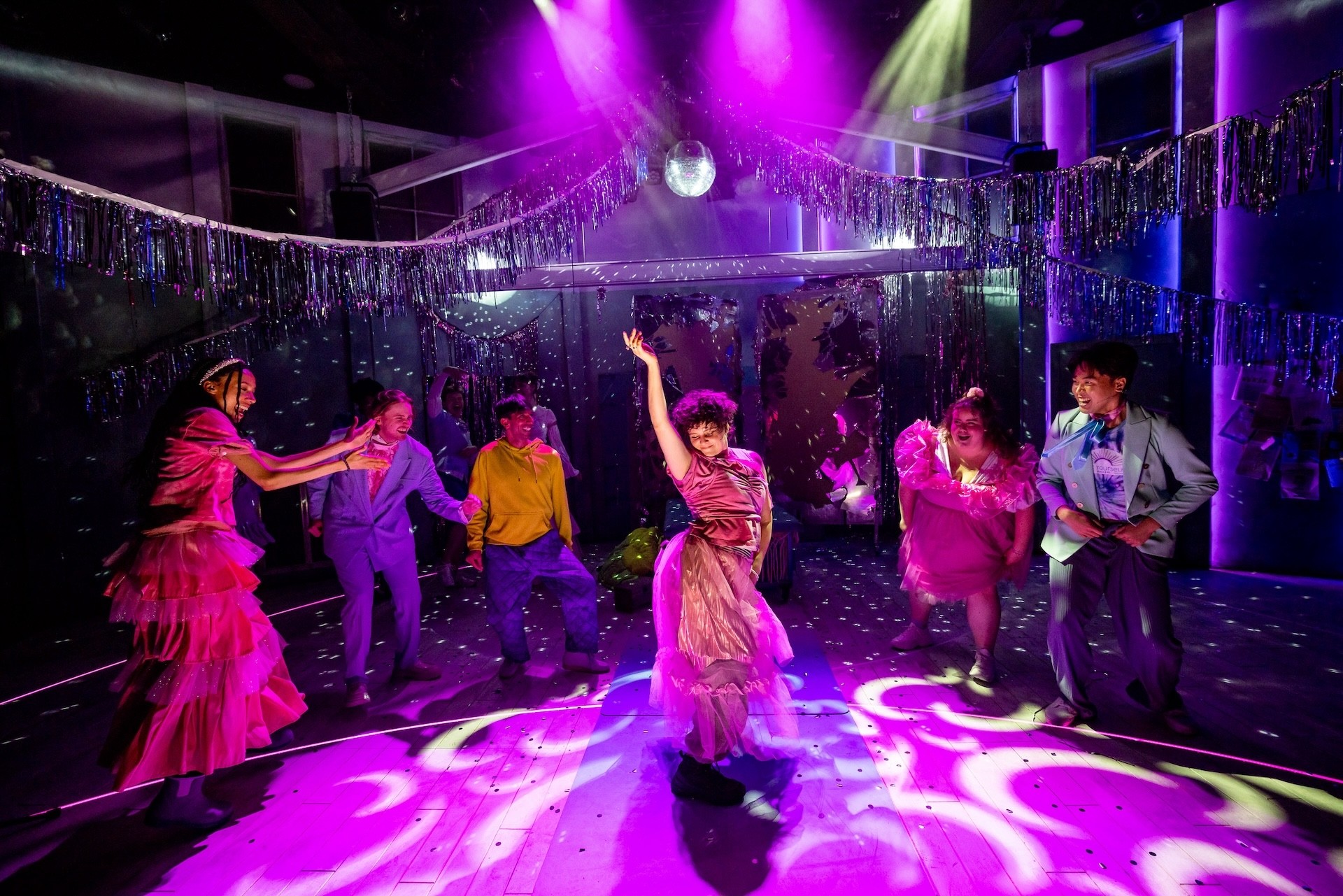 Performers dance under pink and purple lights on a decorated stage with tinsel streamers and a disco ball. A central performer strikes a dramatic pose as the ensemble dances joyfully around them.