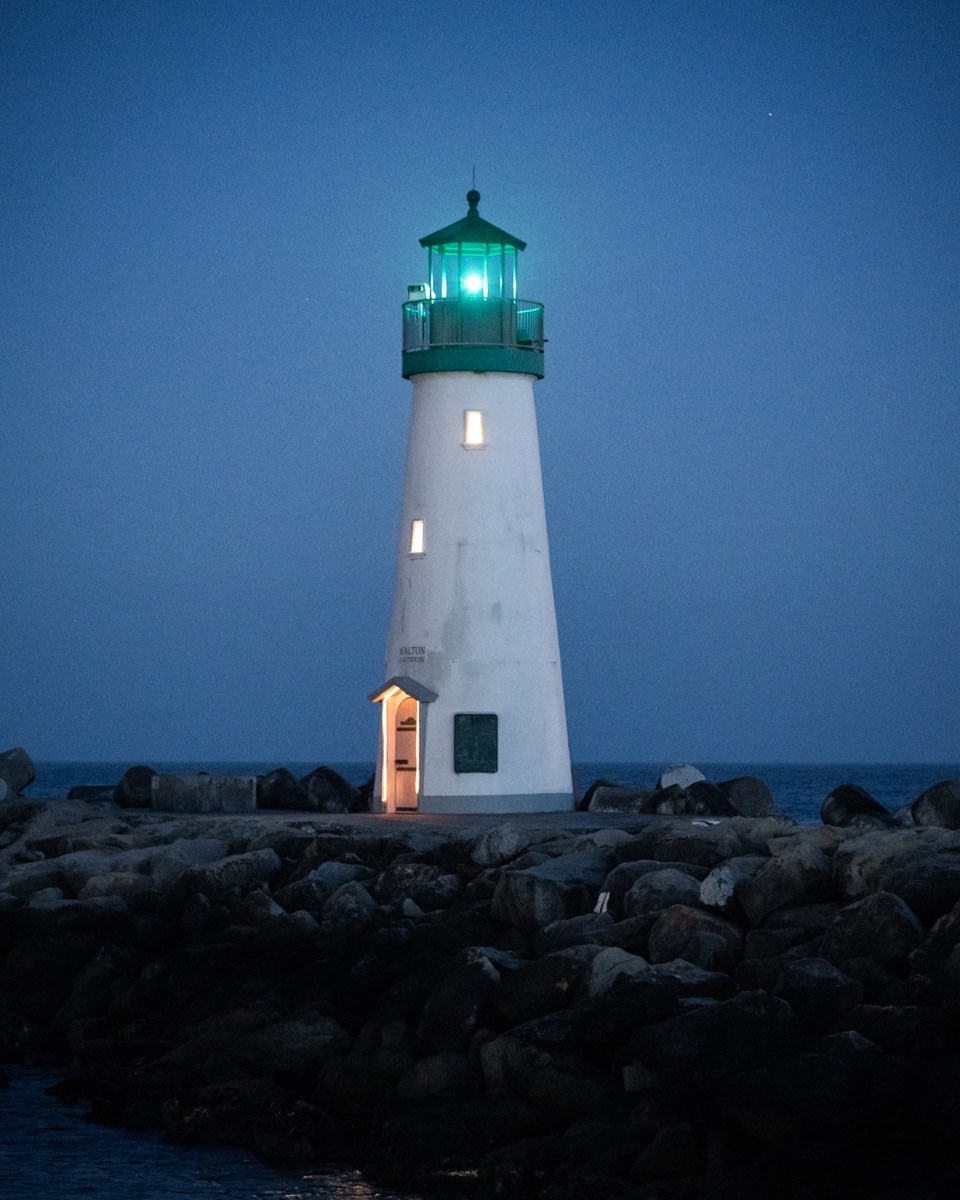 A white lighthouse with a glowing green lantern room stands on a rocky breakwater against a dusky blue sky.