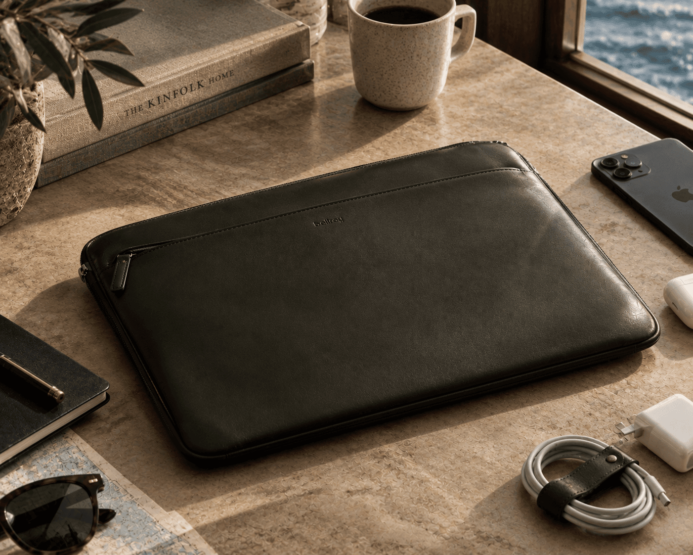 A warm, naturally lit tech accessory flatlay arranged on a beige travertine stone desk next to a window overlooking the ocean. The focal point is a dark charcoal black Bellroy leather laptop sleeve with a zippered front pocket. Styled around the sleeve are essential everyday accessories: a black smartphone, wireless earbuds, a coiled charging cable with a leather strap, a power adapter, a black notebook with a pen, tortoiseshell sunglasses resting on a map, stacked design books, a potted plant, and a ceramic mug filled with dark coffee.