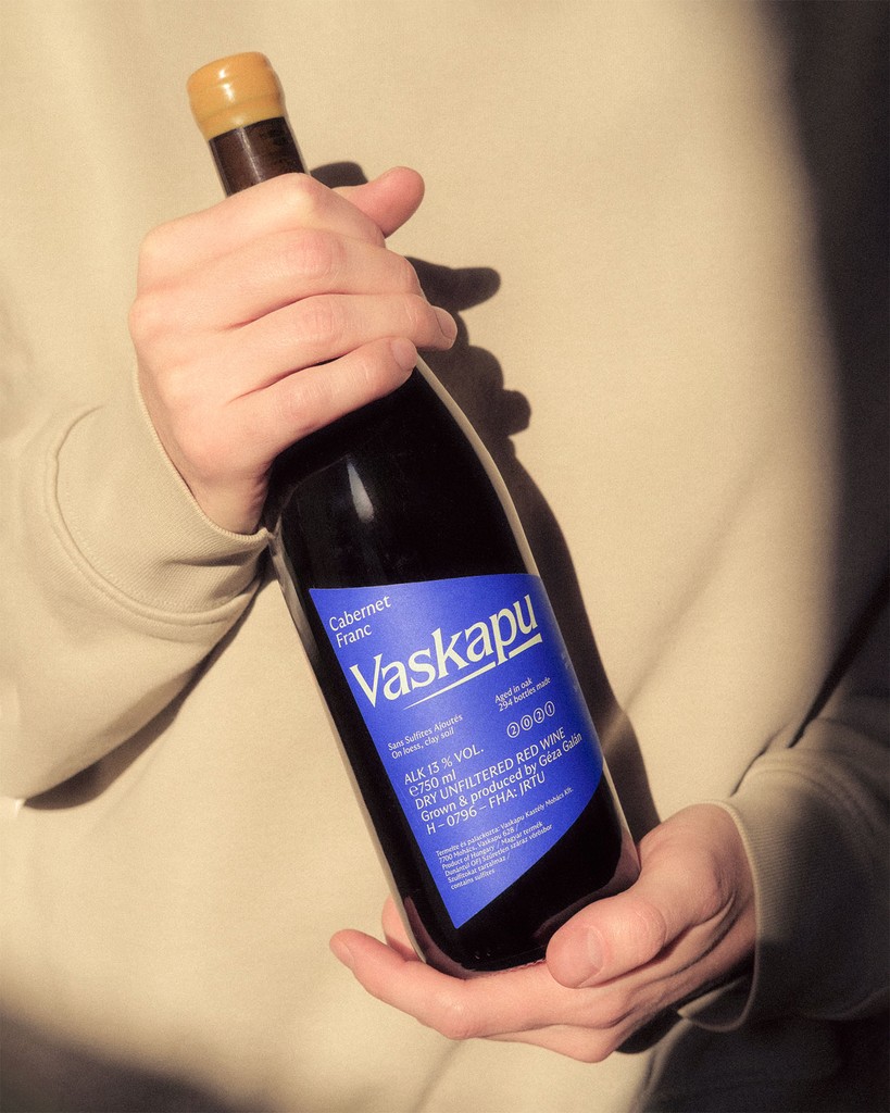 Close-up of a hand holding a Vaskapu Cabernet Franc bottle designed by Marcell Kazsik — minimalist label with deep blue typography, expressing the winery’s natural wine ethos and contemporary Central-European visual language.