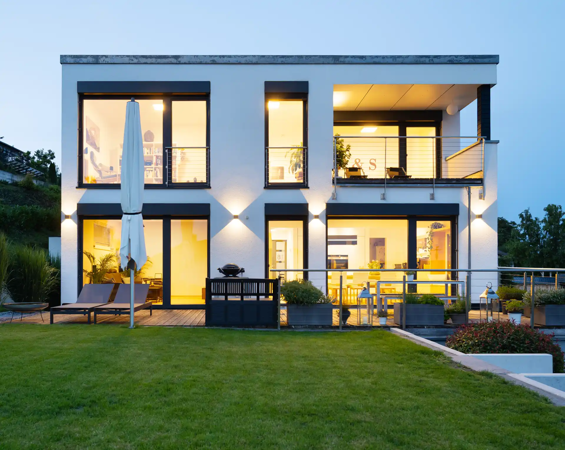Moderne Villa mit großzügigen Glasfronten und beleuchteter Terrasse am Abend, mit gepflegtem Rasen im Vordergrund.