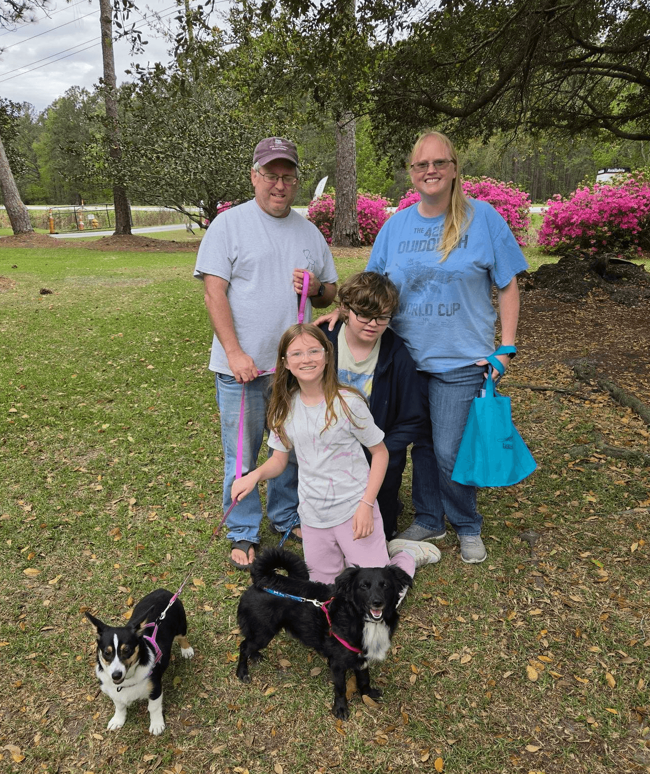 A family stands together outdoors, with two children and two small dogs, surrounded by greenery and blooming flowers.
