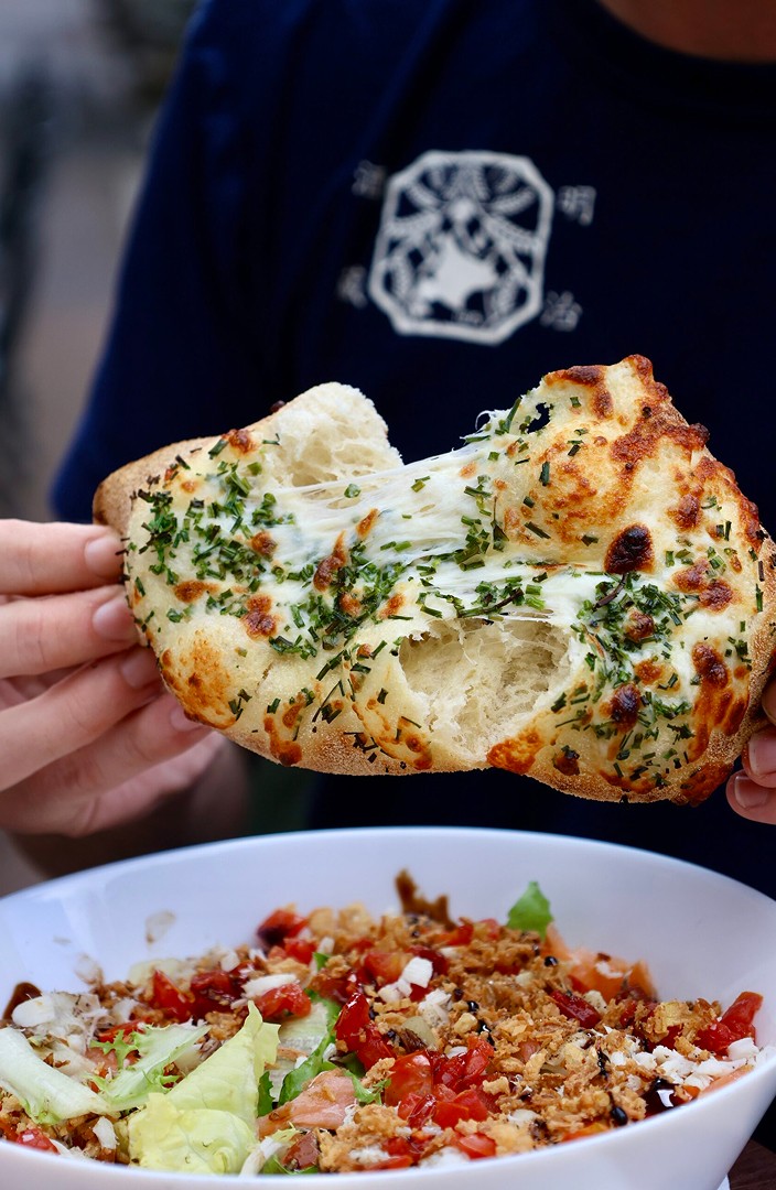 Un pain à l'ail frais et fromagé, garni d'herbes, est tiré, révélant un intérieur de fromage fondant, tenu au-dessus d'un bol blanc rempli d'une salade colorée composée de laitue, de tomates en dés et de garnitures croustillantes.