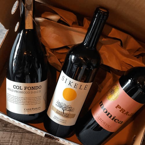Three bottles of Italian wine—Col Fondo, Sikelé, and Pranu Monica—arranged in a box with brown wrapping paper.