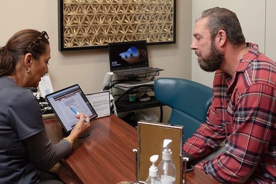 A woman and a man sit at a table, engaging in conversation while looking at a tablet.