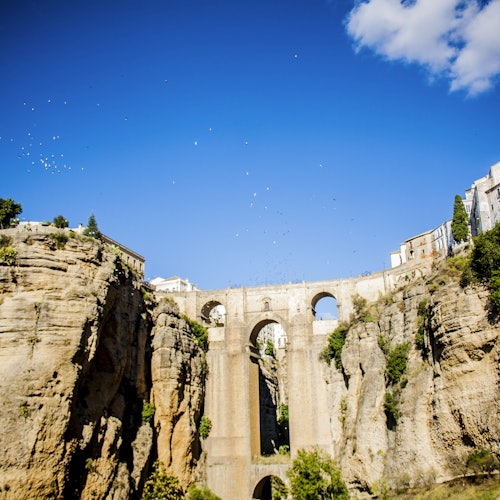 Korkea kivisilta, jossa on useita kaaria, kulkee syvän rotkon yli, taivaalla lentävät linnut ja jyrkänteet ovat vehreydestä vihreinä.