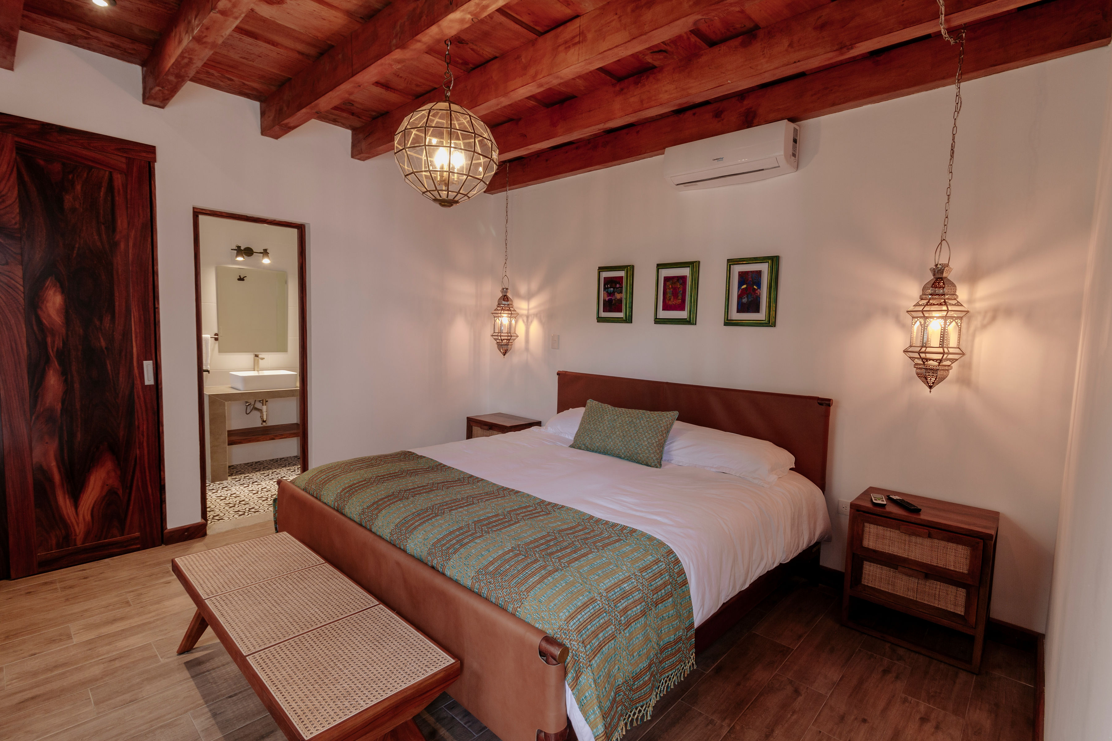 A cozy, rustic bedroom features a large bed with white linens and a green throw blanket, wooden furniture, warm hanging lights, and framed artwork on the walls, creating an inviting ambiance.