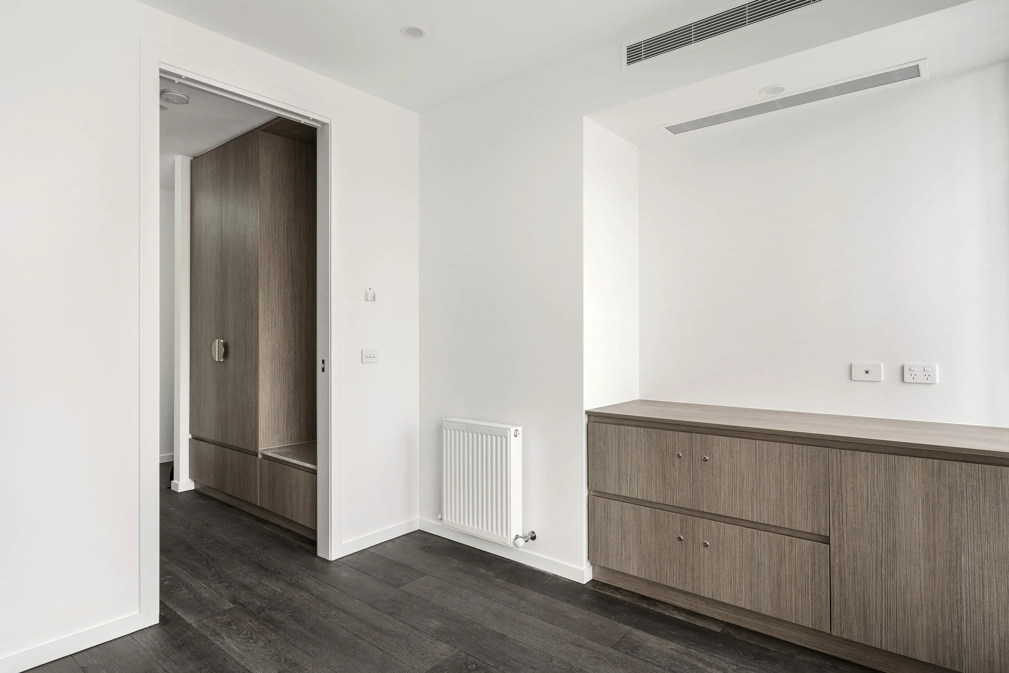 Bedroom with white walls, dark timber floors, built-in oak-toned drawers and a walk-in robe glimpsed through the doorway