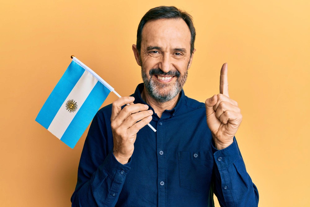 Hombre sonriente mostrando un pulgar hacia arriba y sosteniendo una pequeña bandera de Argentina, con un fondo uniforme de color naranja