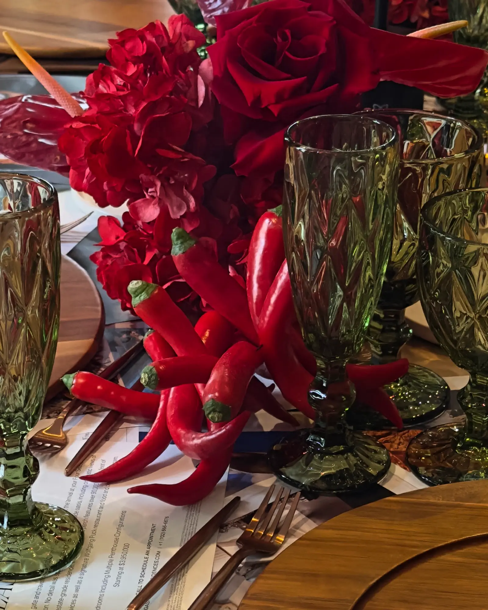Centro de mesa con flores rojas, guindillas y cristalería verde sobre bajoplato de madera y cubertería cobre.