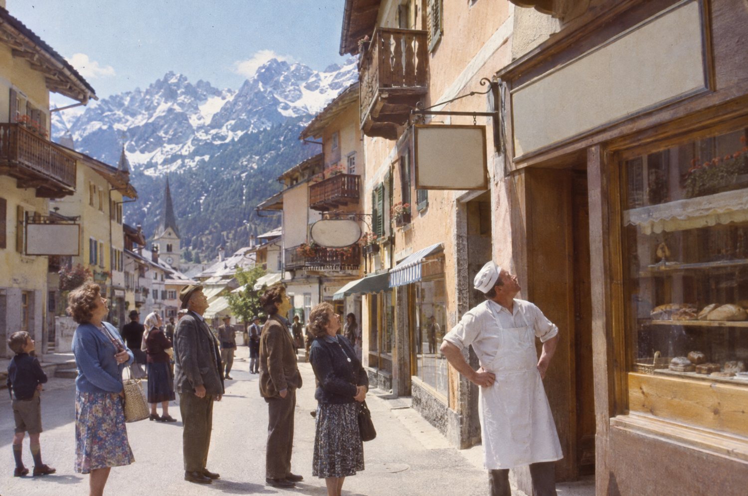 Townspeople and a baker in a white apron look up at blank shop signs on a mountain town's main street