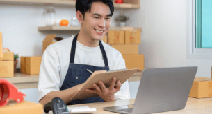 A smiling man looks at a laptop computer while taking notes.