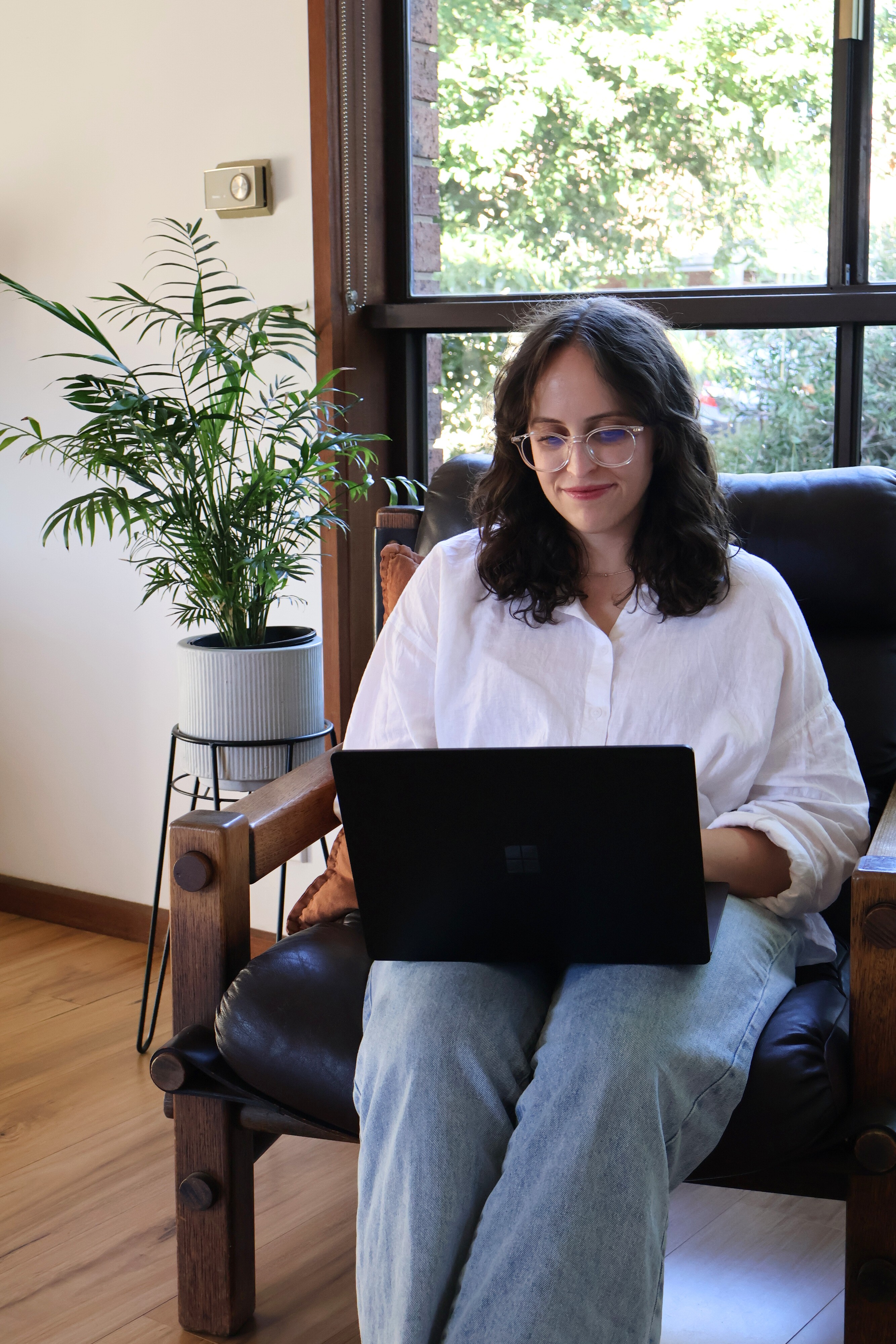 Woman in her late 20s sitting on a brown leather chair in front of a fern plant. She has brown curly hair and is wearing clear-rimmed glasses, a white shirt and pale blue jeans. The woman is typing on a black laptop and smiling at the screen.