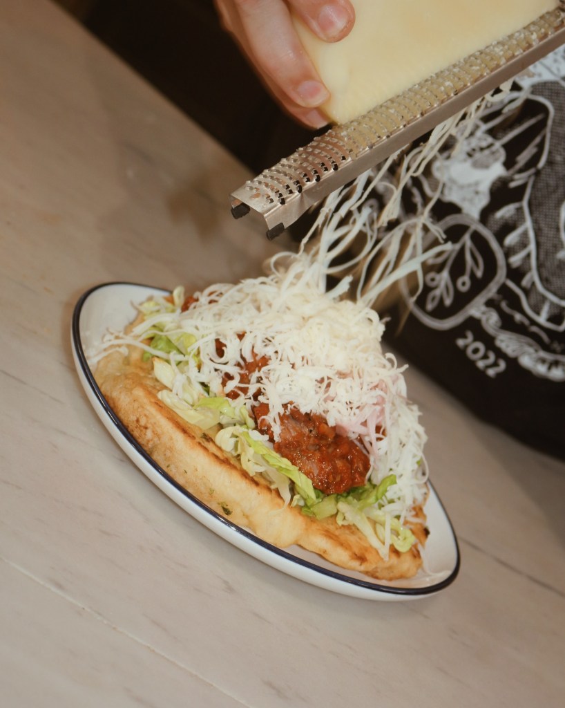 A close-up of a hand grating cheese onto a dish topped with lettuce, meat sauce, and shredded vegetables on a plate.
