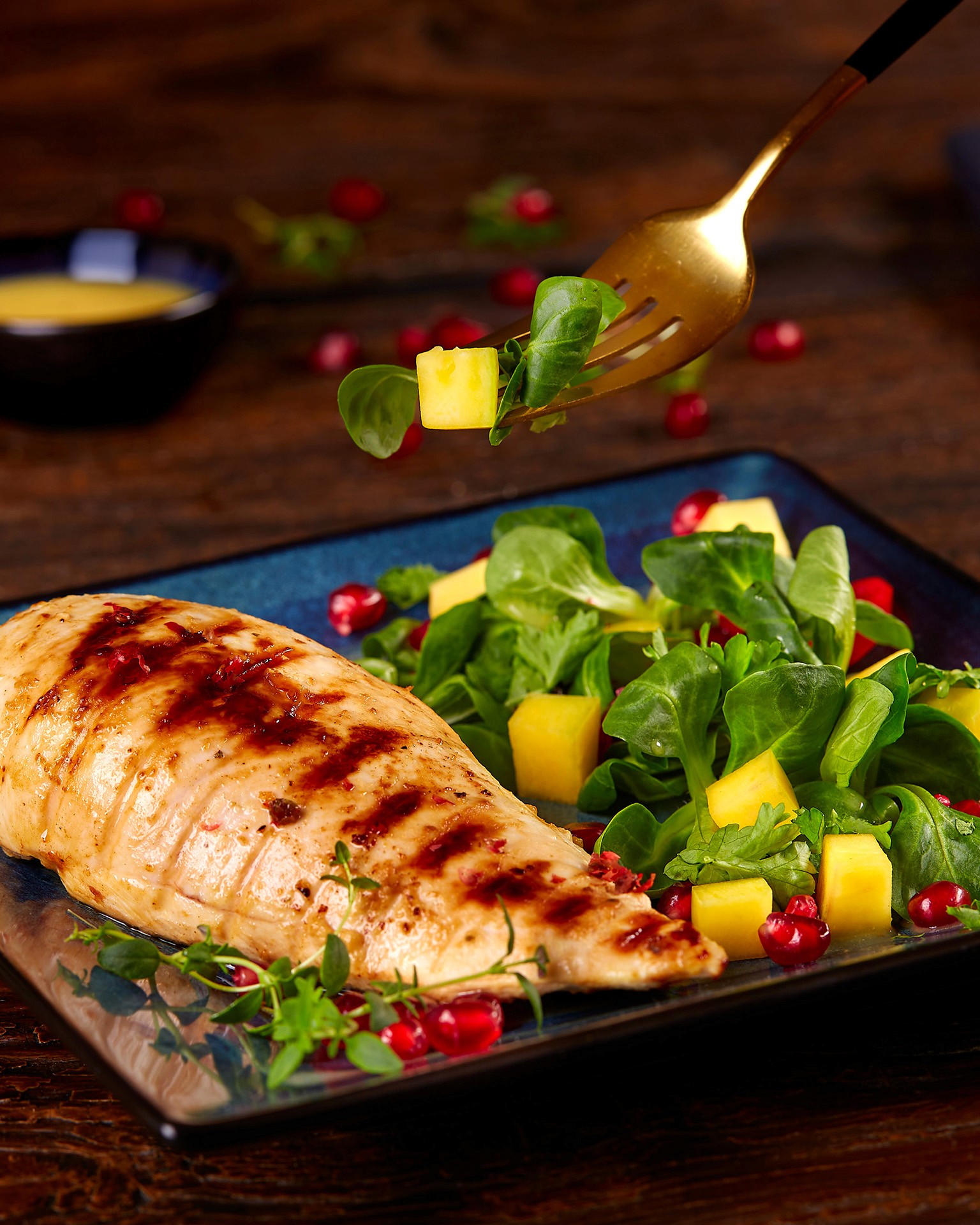 Grilled chicken breast served on a dark square plate with fresh arugula, diced mango, and scattered pomegranate seeds, showcasing a vibrant and healthy meal option.