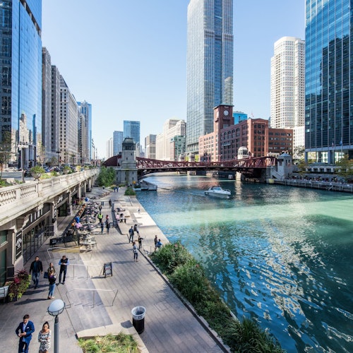 People walking and sitting along the riverwalk with tall buildings and a red bridge in the background on a sunny day.