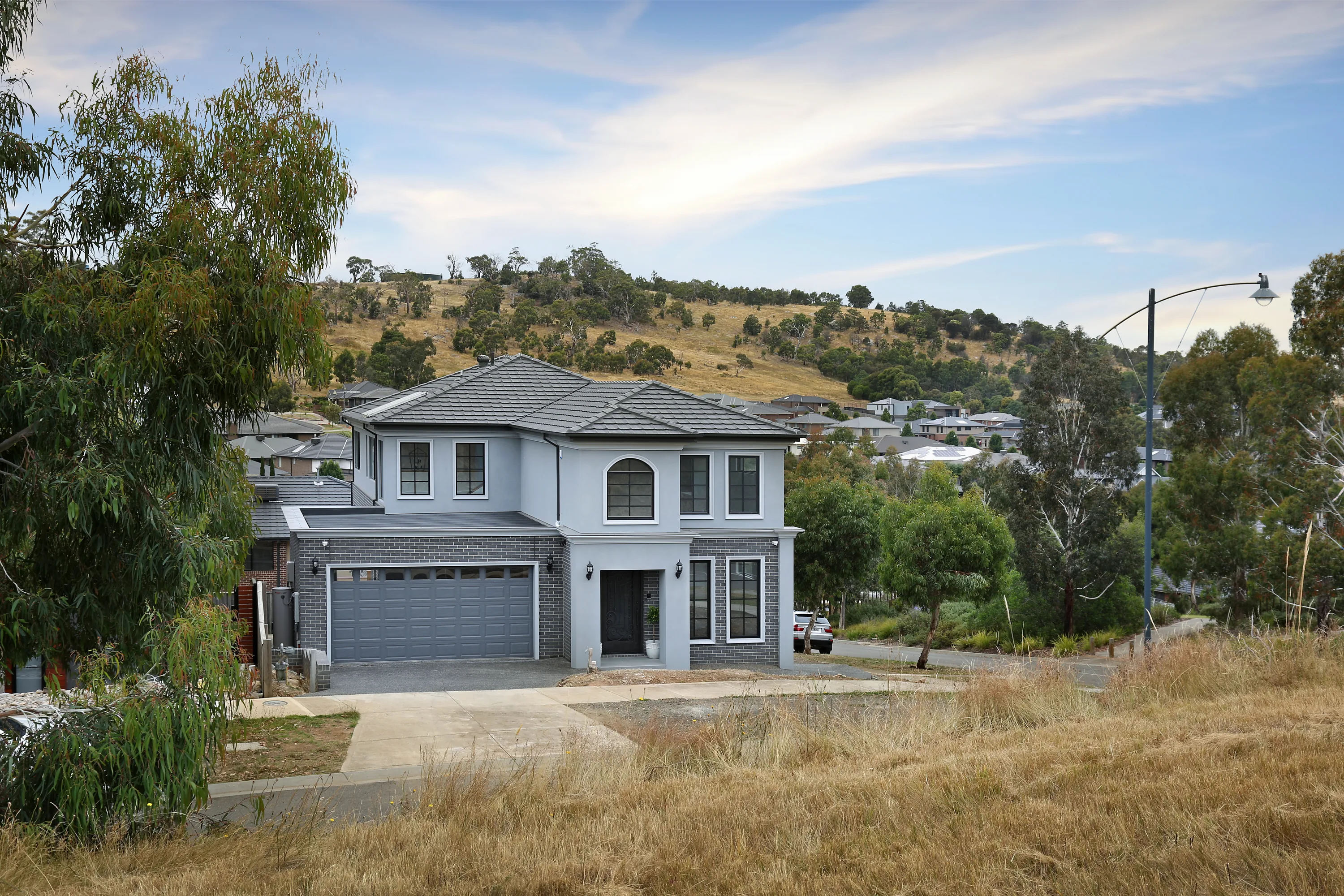 Front facade of South Morang Residence on an elevated leafy block, with soft grey render and face brick, a hipped grey-tiled roof, double garage, arched feature window and a dry grassed hill behind