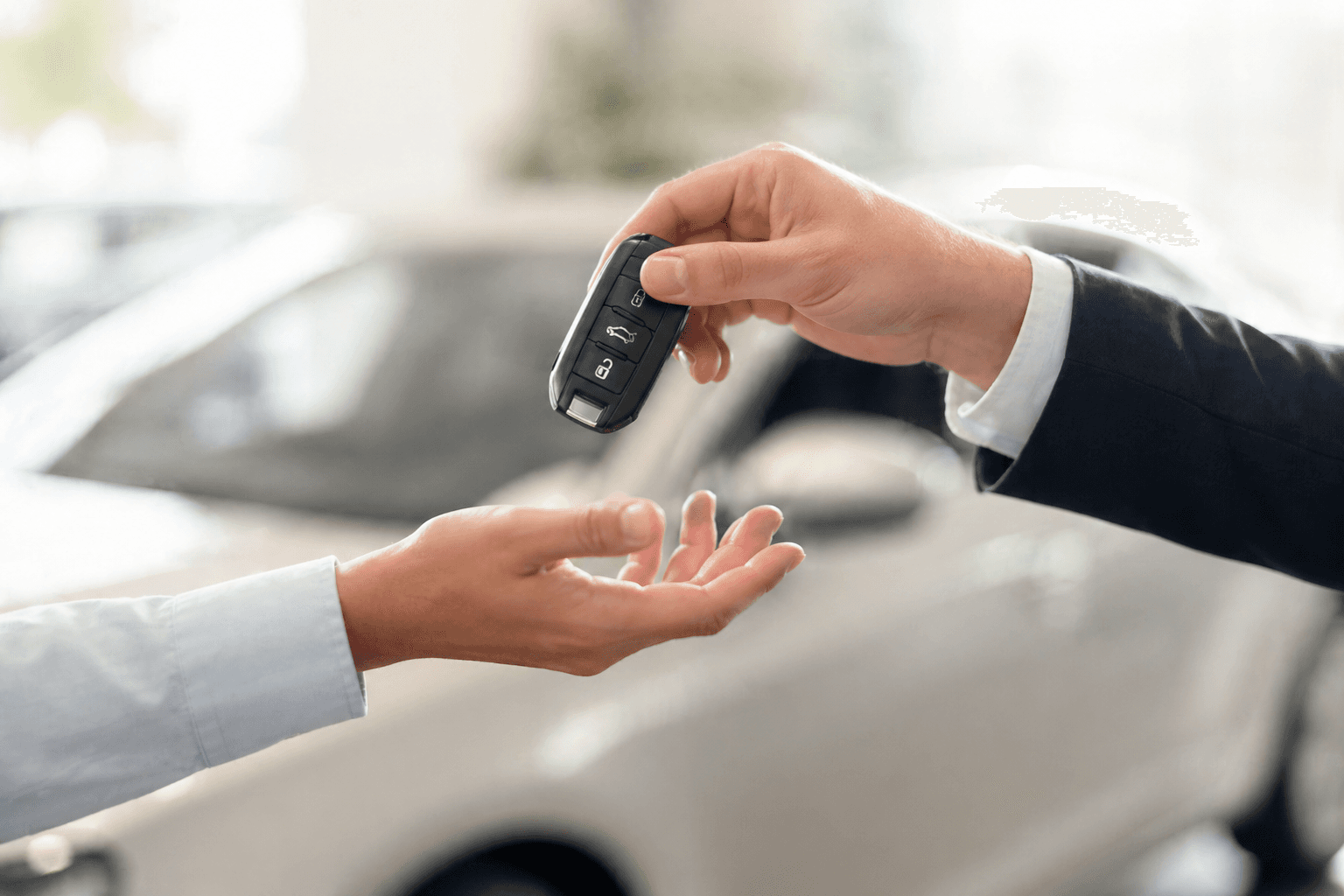 Close-up of one person handing a car key fob to another in a bright car showroom, with a blurred vehicle in the background.