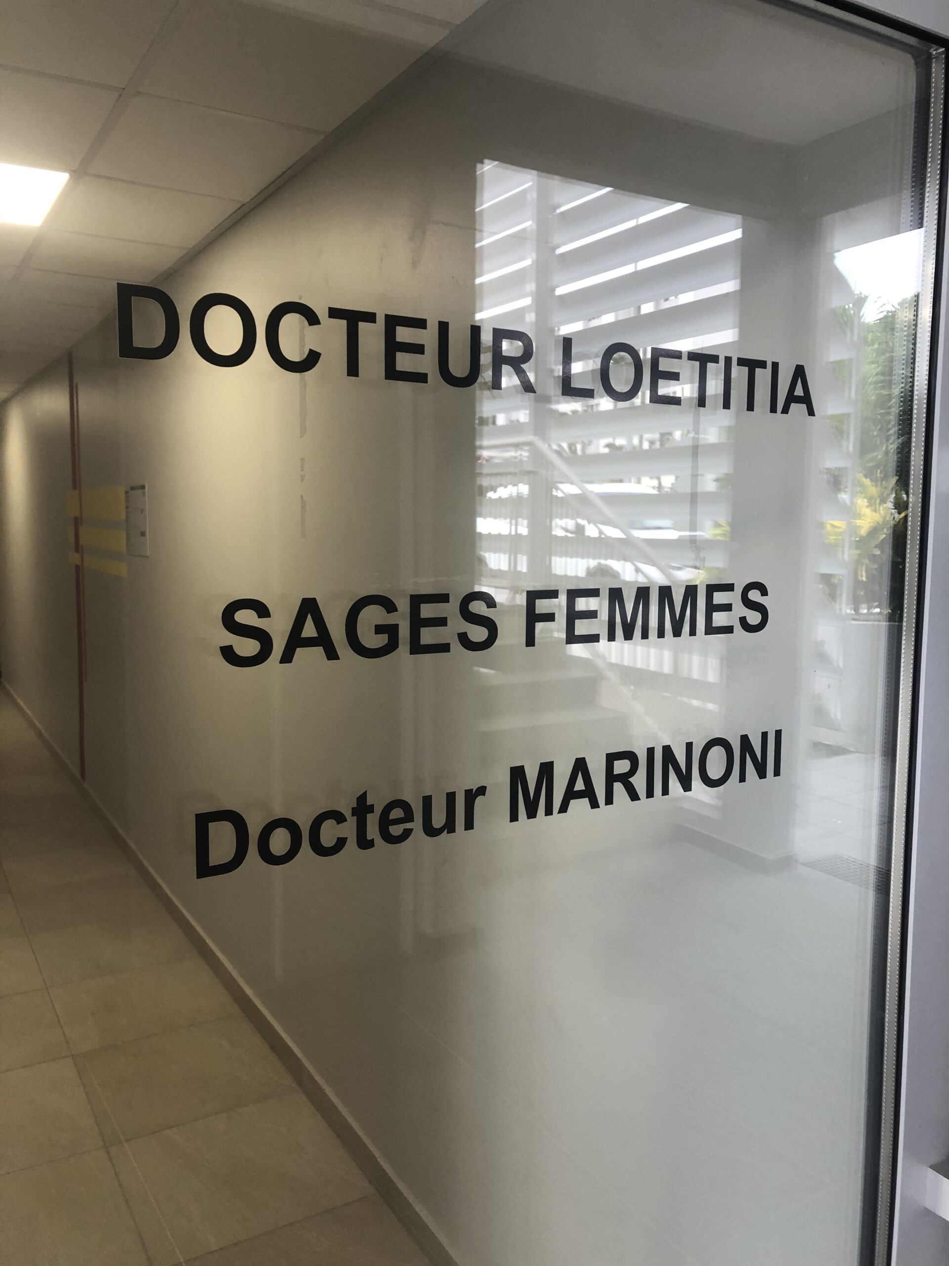 Porte de bureau en verre dépoli avec inscription en noir indiquant des noms et « SAGES FEMMES ». Le couloir est visible sur la gauche, créant une atmosphère professionnelle et clinique.