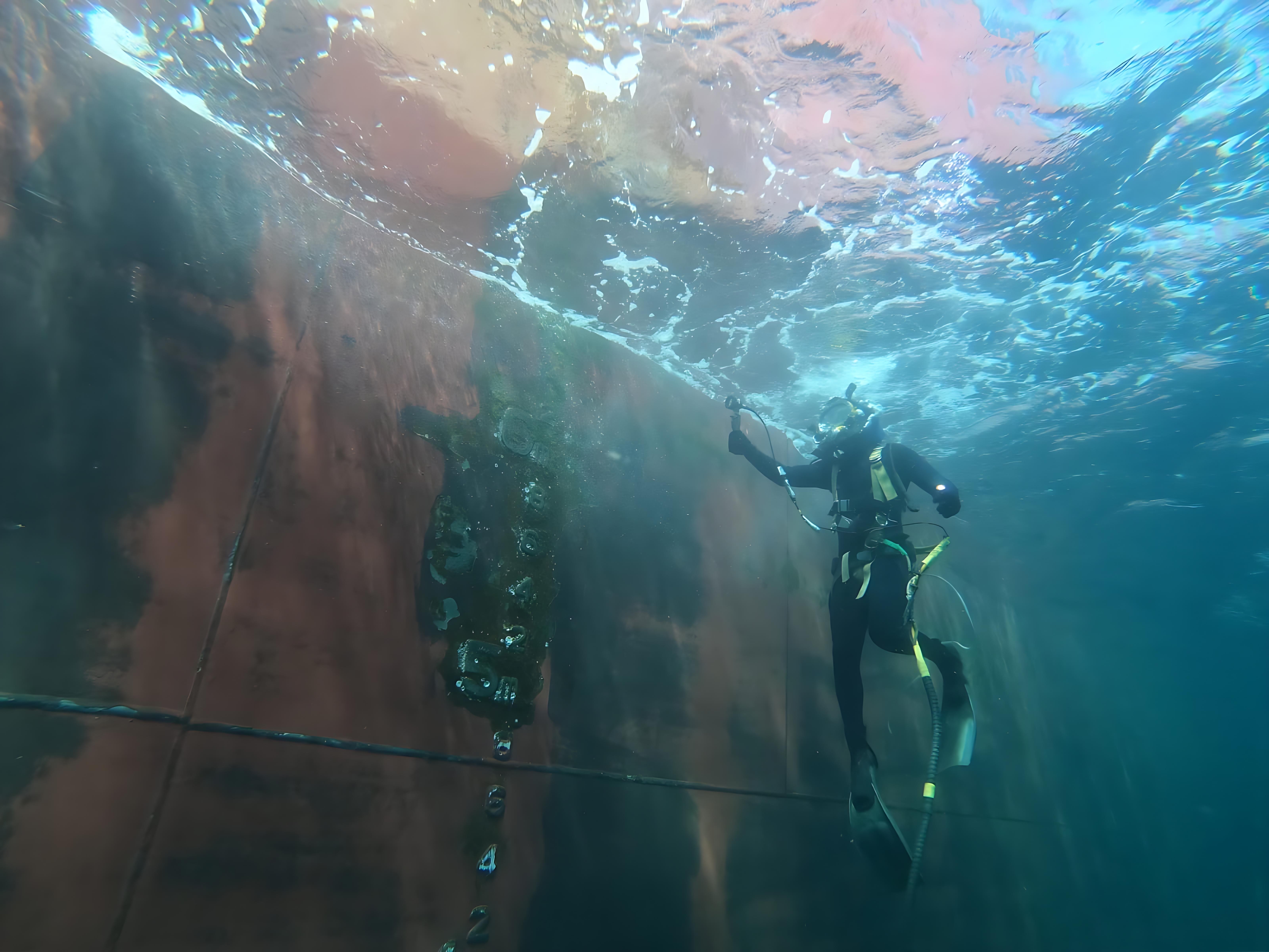 Seachios® Marine Services commercial diver performing close-up underwater hull inspection and spot cleaning on a trading vessel’s shell plating, documenting marine growth and coating condition as part of class-approved underwater survey and hull maintenance program.