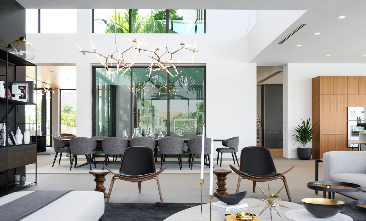 Contemporary luxury living and dining space featuring a sculptural branching chandelier, mid-century modern shell chairs in walnut and black leather, and a long dining table with grey upholstered chairs. Floor-to-ceiling windows bring in natural light and palm tree views, while white walls, light floors, and wooden built-ins create a sleek, modern aesthetic.