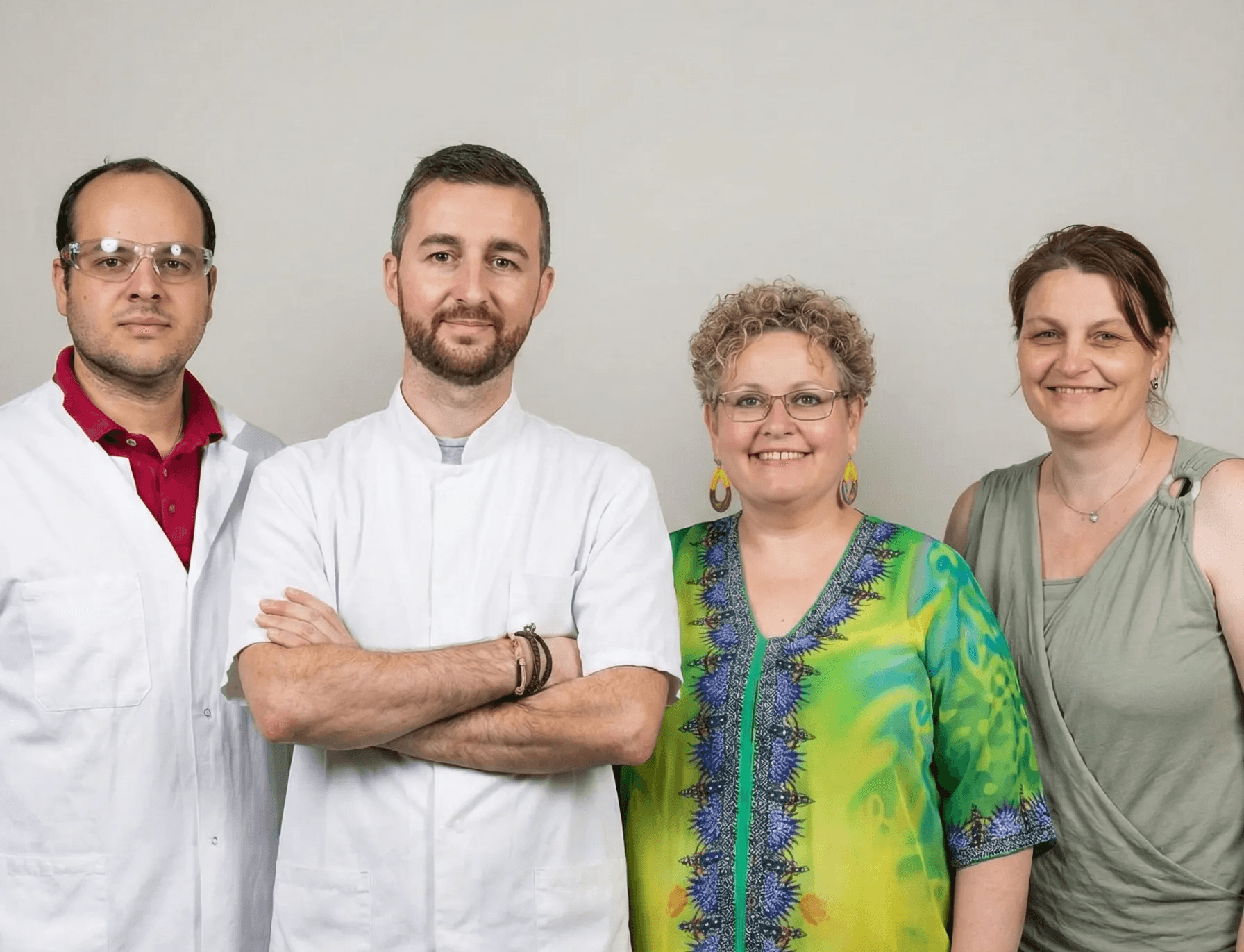 Four people stand against a plain background, with two wearing white lab coats suggesting a professional setting, and two wearing casual colorful clothing, highlighting diversity and teamwork.
