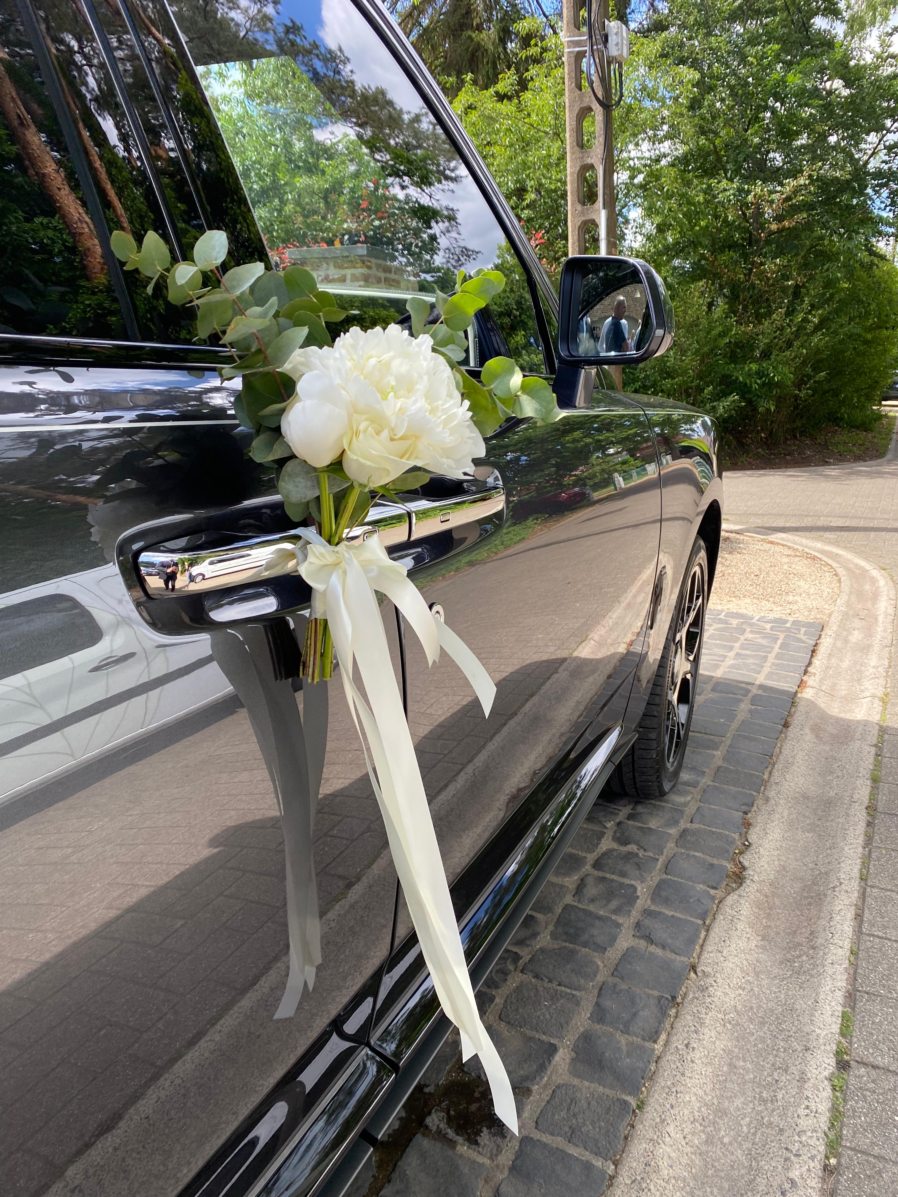 Close-up van de rechterzijkant van een auto, ter hoogte van de achterzijde van de voordeur. Aan de deurklink hangt een bruidsboeket.