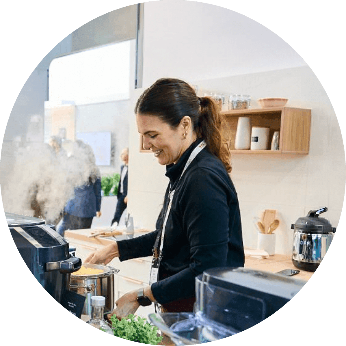 Woman cooks at a stovetop with steam rising from a pan in a modern kitchen, smiling as she prepares food