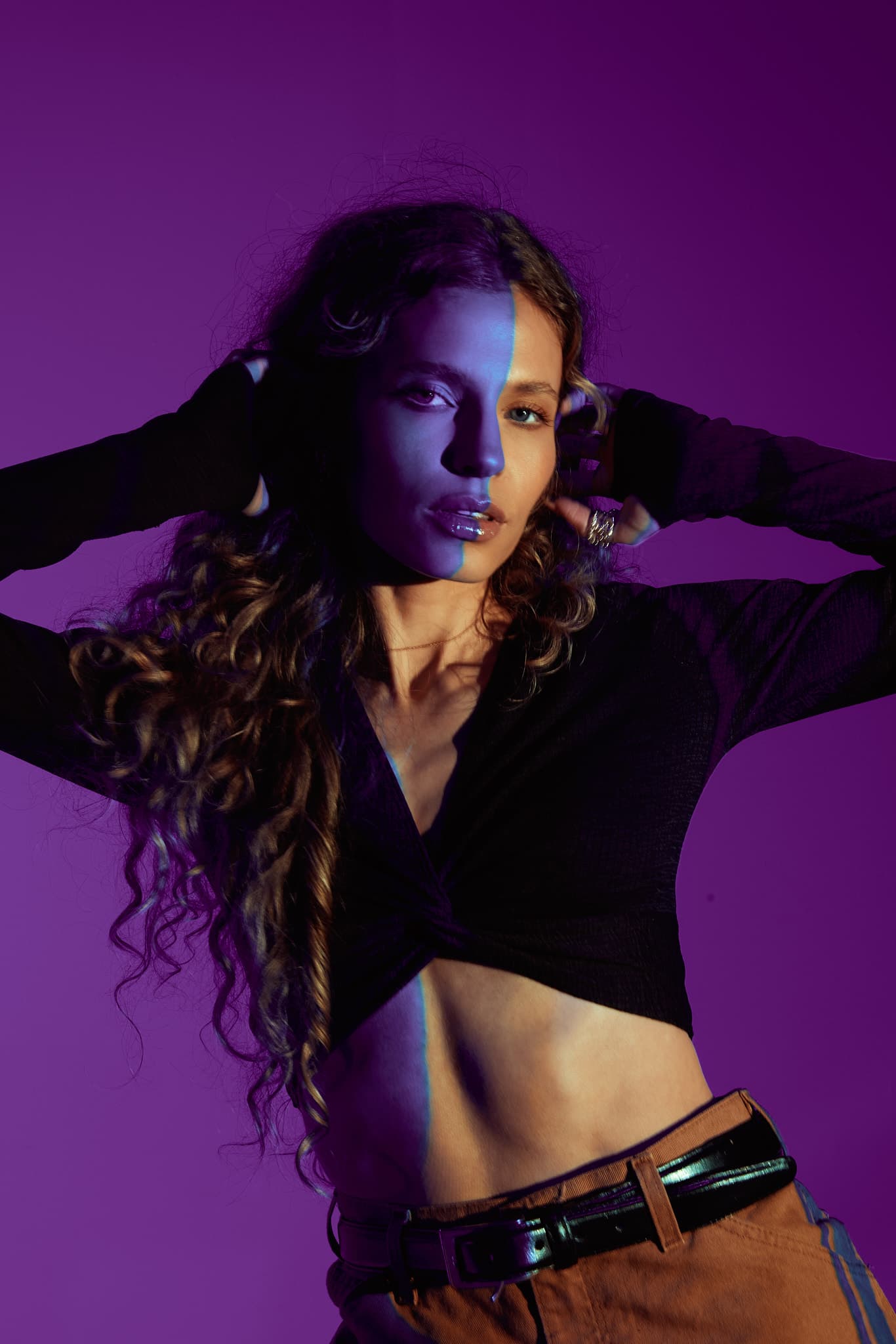 The model poses with her hands on her head in front of a purple background in a natural light studio.