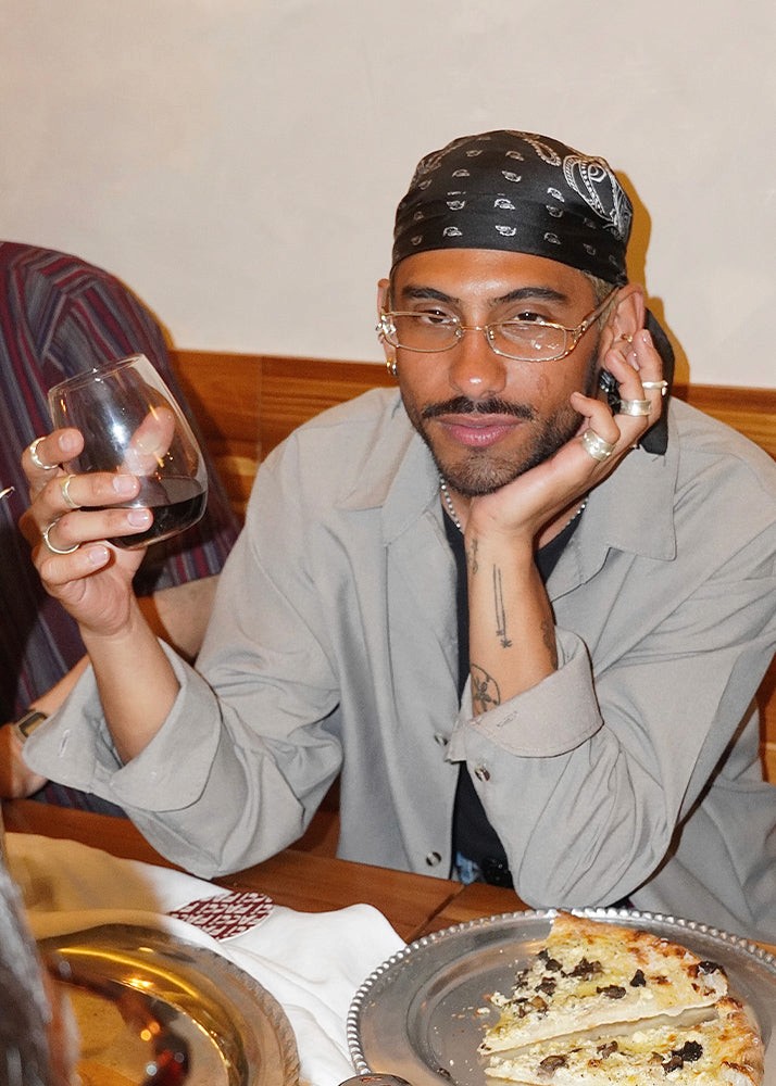 Man wearing silver jewelry sitting at a table with a glass of red wine and a pizza