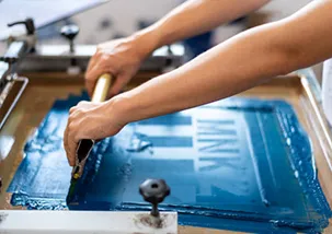 A person is doing screen printing with a 2 hands holding the squeegee with blue ink 