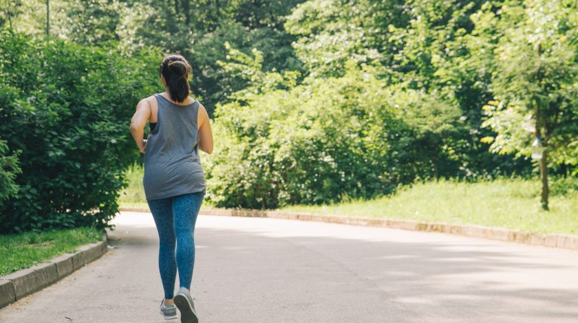 A person in athletic wear is jogging on a paved path surrounded by lush, green trees in a sunny park.
