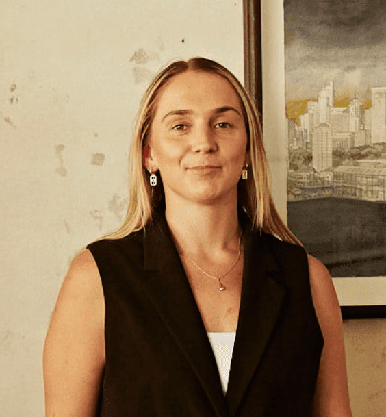 Person wearing a blue puffer jacket and red backpack standing in front of a light blue building on a city street.