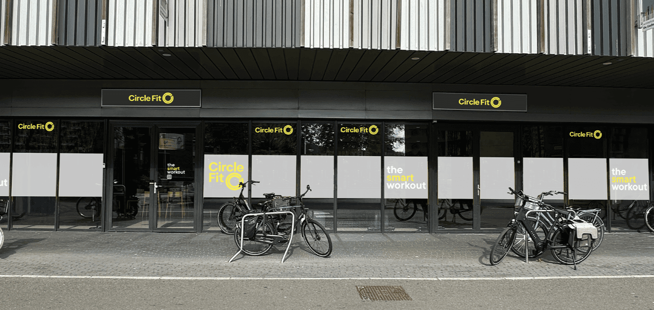 A Circle Fit hub with large glass windows featuring bold yellow signage and people visible indoors. Sidewalk in view.