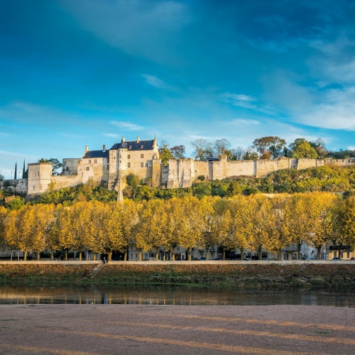 Castillo de piedra histórica con murallas circundantes en la cima de una colina, enmarcado por árboles otoñales y reflejado en un río tranquilo abajo.