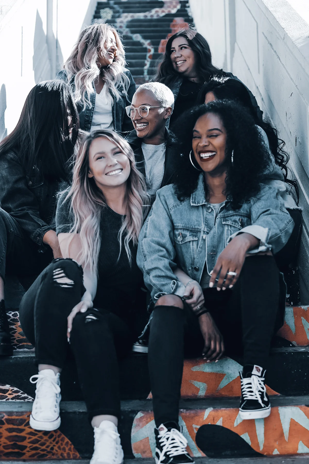 Um grupo diversificado de amigas usando roupas casuais jeans e pretas se senta e ri em uma escadaria grafitada, aproveitando a luz do sol.