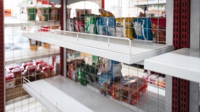 Prateleiras brancas de metal vazias em destaque com outras gôndolas abastecidas ao fundo, em ambiente de supermercado.