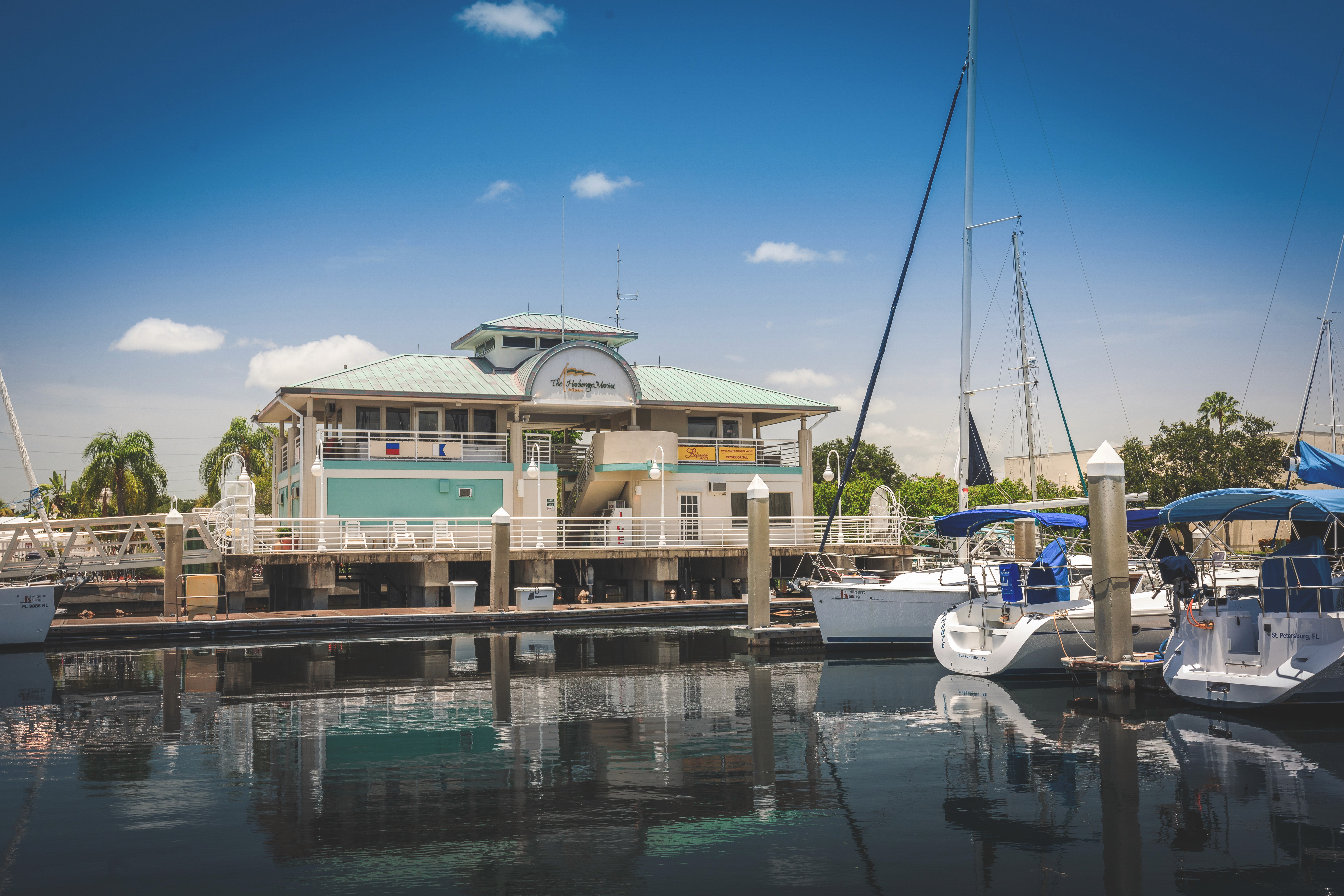 The picturesque Harborage Marina At Bayboro St Pete Florida scene with sailboats moored in calm water, reflecting the clear blue sky and fluffy clouds, with a pastel-colored harbor building in the background.