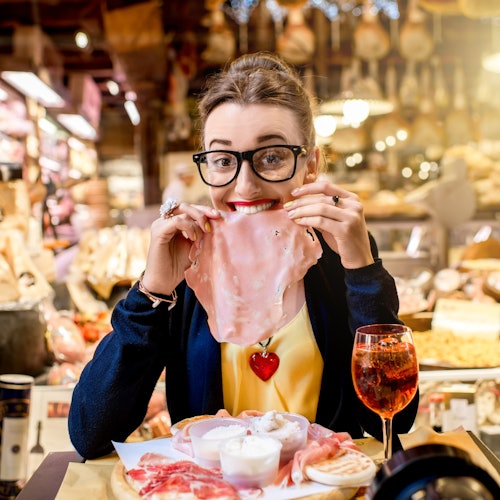 Una donna con gli occhiali tiene una fetta di carne vicino al viso, seduta al bancone di una salumeria con vari tipi di carne e una bevanda rossa.