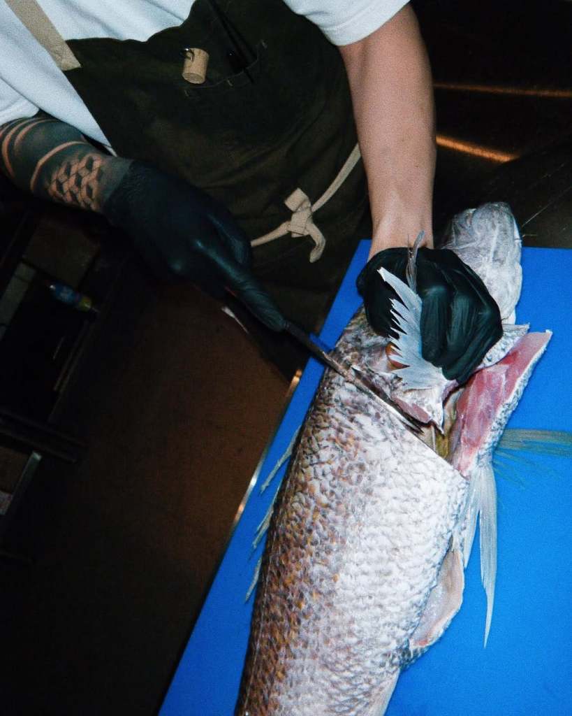 A chef skillfully filleting a large fish on a blue cutting board, wearing gloves and an apron.