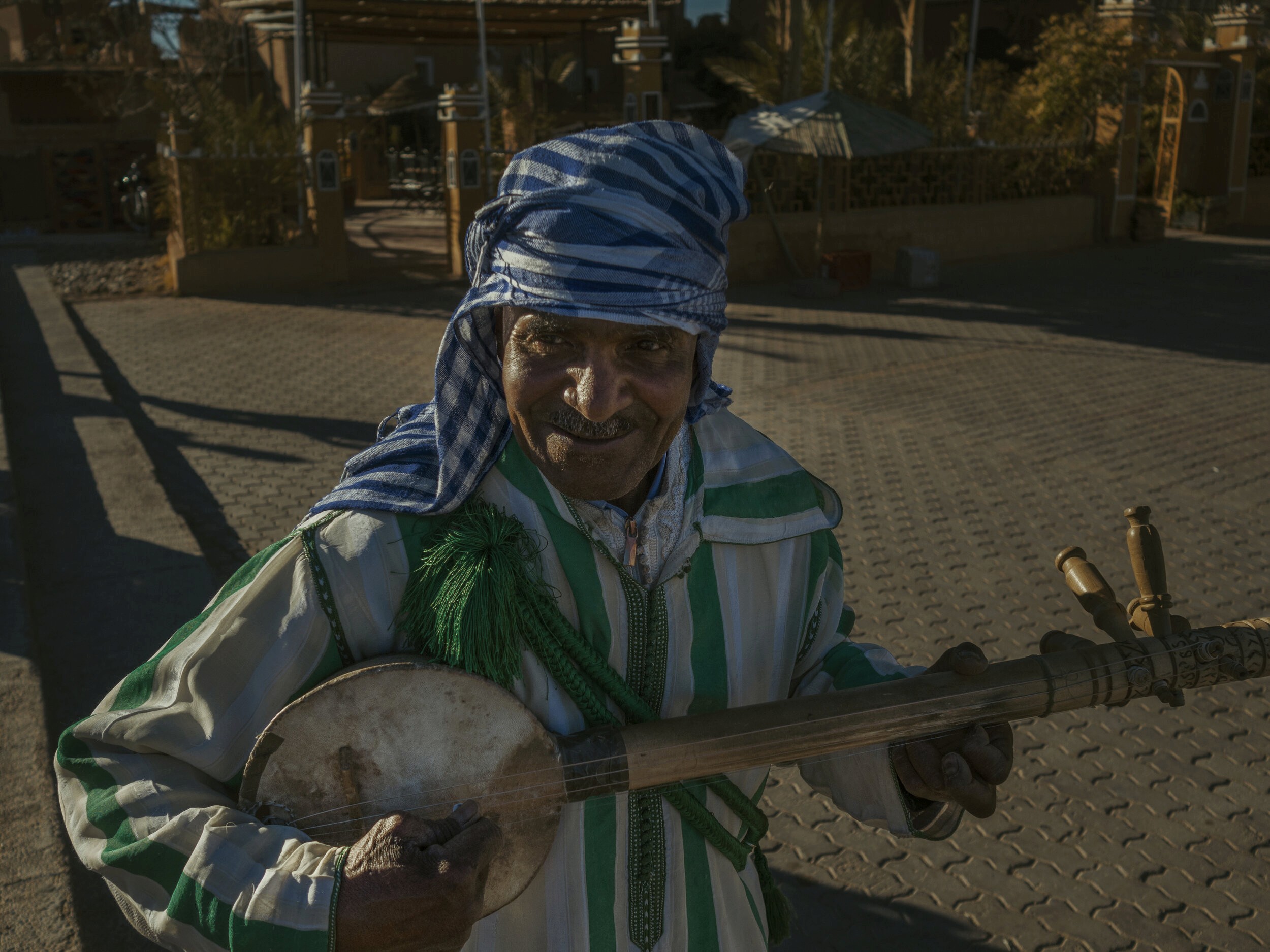 Smiling elderly Moroccan man in a headscarf, holding a traditional instrument in a village square, bathed in warm sunlight.