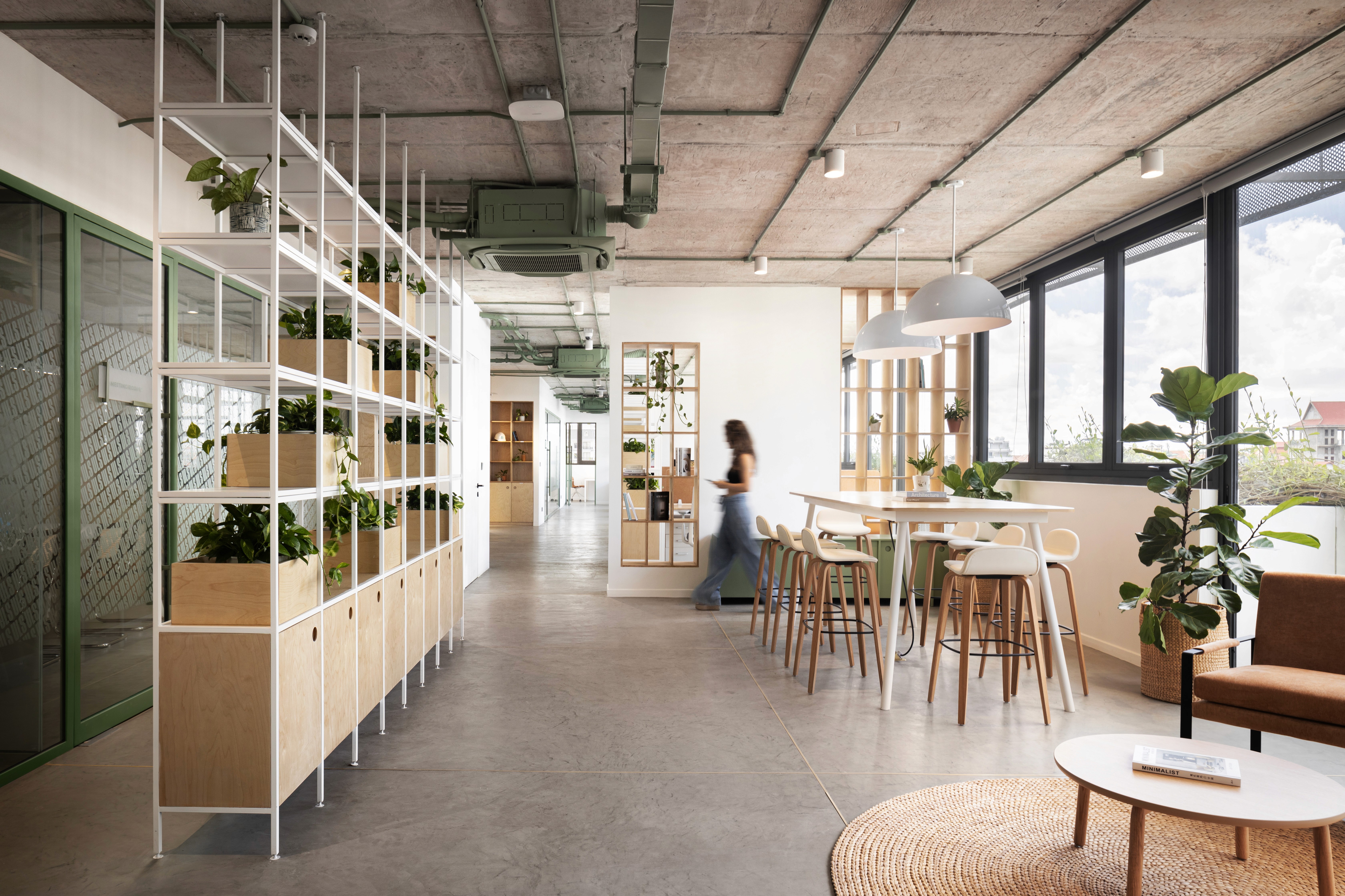 Interior design of the modern reception and entrance area at Urban Living Solutions HQ in Rose Apple Square, Siem Reap, featuring a wooden slatted reception desk with a neon logo, integrated large plants, and an adjacent display area with framed artwork.