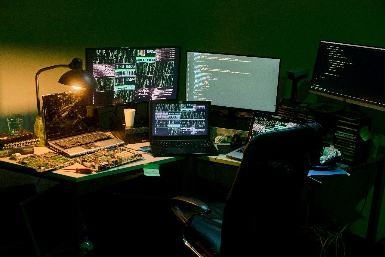 Dimly lit room with multiple computer monitors and laptops displaying code and data. Electronic equipment is scattered on the desk, conveying a busy, tech-focused atmosphere.