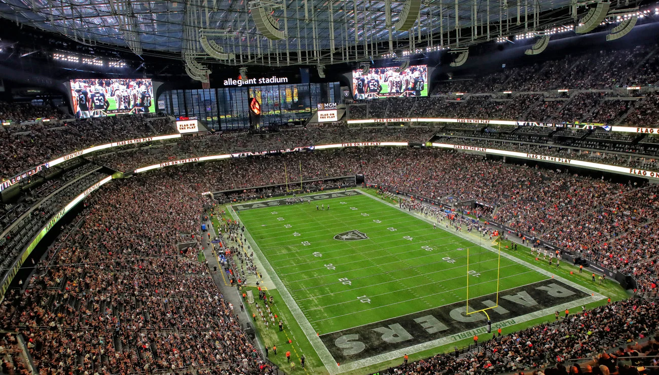 Wide view inside Allegiant Stadium packed with fans during a Raiders home game. The bright green field sits below large video boards and a glass wall, with “Raiders” visible in the end zone.